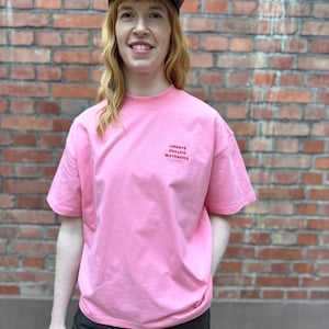 Könnte beinhalten: Ein rosa T-Shirt mit den Worten "LIBERTÉ ÉGALITÉ MATERNITÉ" in roter Schrift. Das Shirt wird mit einer schwarzen Hose und einer grau-schwarzen Kappe kombiniert. Der Hintergrund ist eine rote Backsteinmauer.