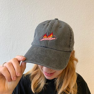 Könnte beinhalten: Eine anthrazitfarbene Baseballkappe mit gebogenem Schirm. Die Kappe hat ein gesticktes Design eines rosa BHs in Flammen. Die Kappe besteht aus einem weichen, abgenutzten Stoff.
