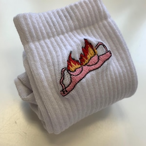 Könnte beinhalten: Weiße gerippte Socken mit einem gestickten Design eines brennenden rosa BHs. Die Flammen sind orange und gelb. Die Socken sind gefaltet und scheinen aus einem weichen, bequemen Material zu sein.