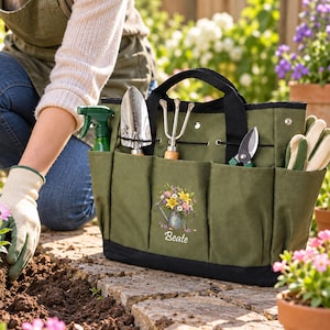 May include: An olive green canvas garden tool bag with black trim and handles. The bag features multiple exterior pockets containing a green spray bottle, a metal trowel, a hand rake, pruning shears, and gardening gloves. A floral graphic and the name Beate are printed on the front.