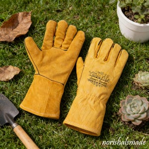 以下が含まれることがあります： 黒いステッチが入った黄色の革製ガーデニンググローブのペア。片方の手袋には「Susan's Garden」の文字がプリントされています。手袋は緑の草の上にあり、小さなシャベルと植物が背景に見えます。