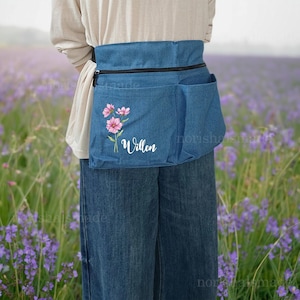 May include: A denim blue waist apron with multiple pockets and a black zipper. The apron features a floral design with pink flowers and the name "Willen" in white script. The apron is worn around the waist.