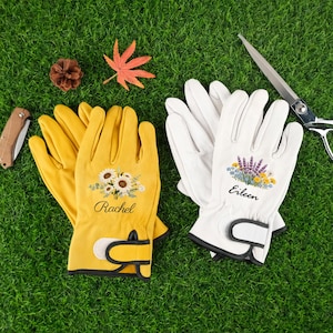 以下が含まれることがあります： 緑の芝生の上に2組のガーデニンググローブ。片方はヒマワリのデザインと「レイチェル」という名前が入った黄色。もう片方は花の模様と「アイリーン」という名前が入った白。ハサミ、ナイフ、松ぼっくり、葉も写っています。