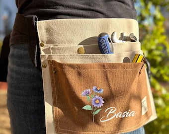 May include: A beige and brown canvas tool belt with multiple pockets. The front pocket features a floral design and the name "Basia" in white script. Tools, including a blue handled screwdriver, are visible in the pockets.