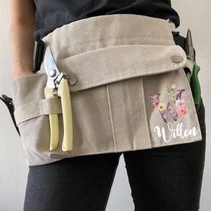 以下が含まれることがあります： ベージュ色のキャンバスガーデニングツールベルト。複数のポケットとループ付き。剪定ばさみなどのツールが含まれています。「W」のイニシャルと「Willen」の名前が花柄でデザインされています。