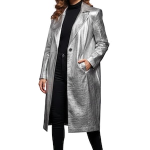 Könnte beinhalten: Ein silberner, metallischer langer Mantel mit Reverskragen und einem Ein-Knopf-Verschluss. Der Mantel hat lange Ärmel und Taschen. Die Person trägt einen schwarzen Rollkragenpullover und eine schwarze Hose.