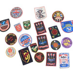 Könnte beinhalten: Eine Sammlung farbenfroher, gestickter Aufnäher mit verschiedenen Designs und Texten. Die Aufnäher enthalten Logos für Dart-Turniere, Organisationen und Marken. Die Aufnäher haben verschiedene Formen, darunter Kreise, Schilde und Rechtecke.