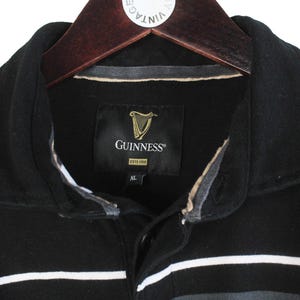 May include: Close-up of a black and gray striped polo shirt with a black collar. The shirt features the Guinness logo on a black tag. The shirt is hanging on a wooden hanger.