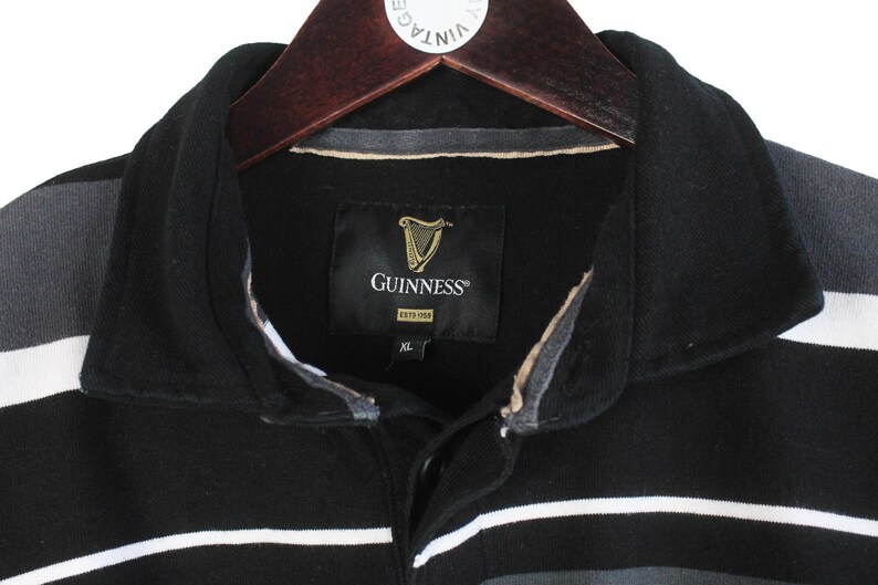 May include: Close-up of a black and gray striped polo shirt with a black collar. The shirt features the Guinness logo on a black tag. The shirt is hanging on a wooden hanger.