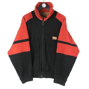 Könnte beinhalten: Eine schwarz-rote Fleecejacke mit Reißverschluss, hohem Kragen, langen Ärmeln mit horizontalen roten Streifen und einem kleinen rechteckigen Aufnäher auf der Brust mit dem Text Think Pink. Das Kleidungsstück hat einen zentralen Reißverschluss.