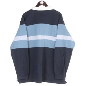 May include: A long-sleeved rugby shirt with a white collar and a color block design. The shirt features horizontal stripes in navy blue, light blue, and white. The shirt is hanging on a wooden hanger.