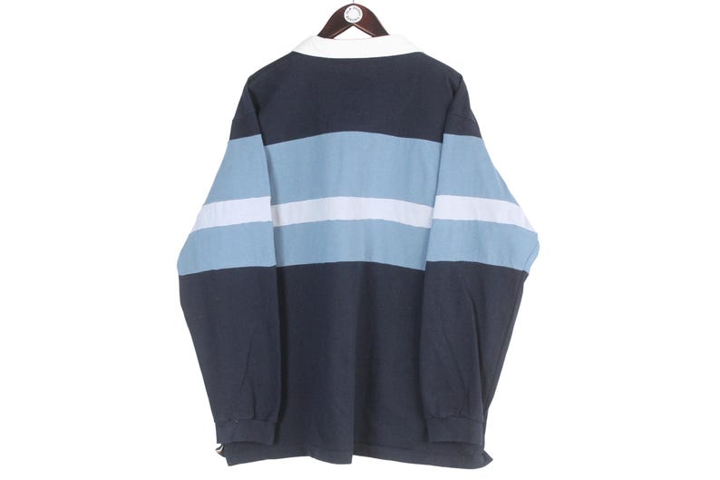 May include: A long-sleeved rugby shirt with a white collar and a color block design. The shirt features horizontal stripes in navy blue, light blue, and white. The shirt is hanging on a wooden hanger.