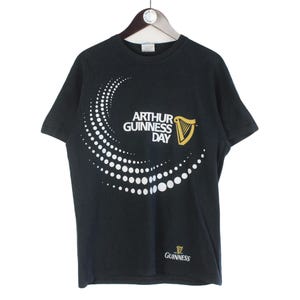 Pu&ograve; includere: T-shirt nera con la scritta "ARTHUR GUINNESS DAY" in bianco, un disegno a pois bianchi e un logo dell'arpa dorata. Il logo Guinness &egrave; in basso.