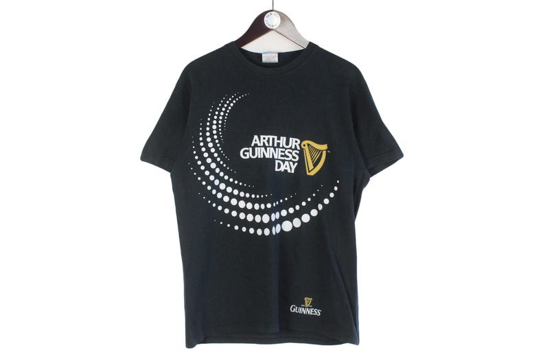 Pu&ograve; includere: T-shirt nera con la scritta "ARTHUR GUINNESS DAY" in bianco, un disegno a pois bianchi e un logo dell'arpa dorata. Il logo Guinness &egrave; in basso.