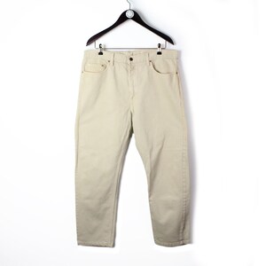 May include: A pair of beige denim jeans featuring a classic five-pocket design, a button fly closure, and a waistband with belt loops. The trousers are constructed from a solid light-coloured cotton fabric and are displayed hanging on a black hanger.