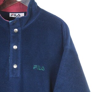 K&ouml;nnte beinhalten: Ein marineblauer Fleece-Pullover mit Knopfleiste und kontrastierendem bordeauxfarbenem Kragen. Das Fila-Logo ist gr&uuml;n auf die Brust gestickt. Das Kleidungsst&uuml;ck hat einen hohen Ausschnitt und drei silberne Kn&ouml;pfe.