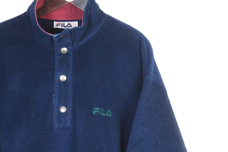 K&ouml;nnte beinhalten: Ein marineblauer Fleece-Pullover mit Knopfleiste und kontrastierendem bordeauxfarbenem Kragen. Das Fila-Logo ist gr&uuml;n auf die Brust gestickt. Das Kleidungsst&uuml;ck hat einen hohen Ausschnitt und drei silberne Kn&ouml;pfe.