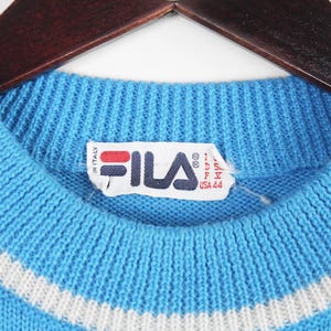 Puede incluir: Primer plano de un su&eacute;ter de punto azul claro con un detalle de rayas blancas en el cuello. El su&eacute;ter tiene un cuello acanalado y una etiqueta con el logotipo de Fila y texto. El su&eacute;ter est&aacute; colgado en una percha de madera.