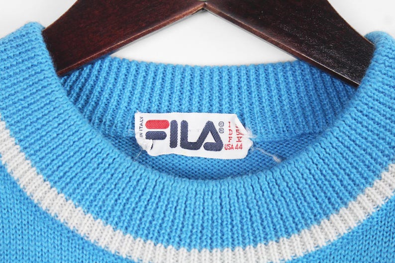 Puede incluir: Primer plano de un su&eacute;ter de punto azul claro con un detalle de rayas blancas en el cuello. El su&eacute;ter tiene un cuello acanalado y una etiqueta con el logotipo de Fila y texto. El su&eacute;ter est&aacute; colgado en una percha de madera.