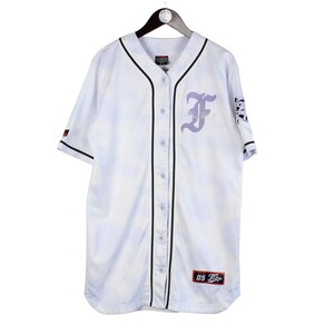 Può includere: Maglia da baseball bianca con finiture nere e chiusura con bottoni. Presenta una "F" viola chiaro sul petto e una toppa con "05" in basso. La maglia è appesa a una gruccia nera.