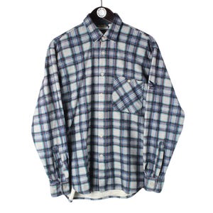 Puede incluir: Una camisa de manga larga con botones y un patrón de cuadros en tonos gris, azul marino y blanco. La camisa incluye un bolsillo en el pecho en el lado izquierdo, un cuello estándar y puños abotonados, con un pequeño logotipo oscuro en la manga.