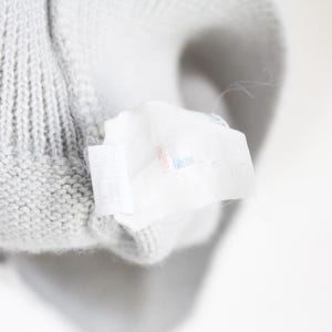 Puede incluir: Primer plano de una tela de punto gris claro con una etiqueta blanca adjunta. La etiqueta parece tener texto y detalles de color. La tela tiene un patr&oacute;n texturizado y acanalado.