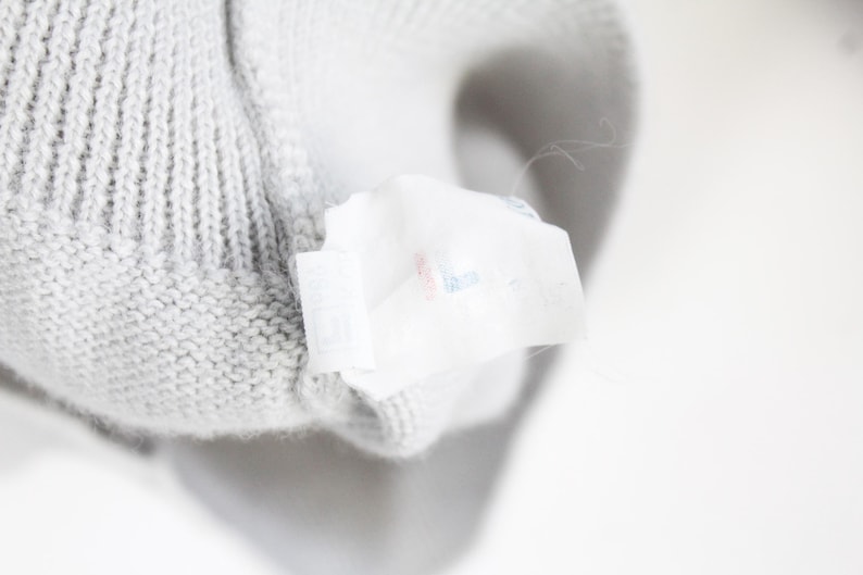 Puede incluir: Primer plano de una tela de punto gris claro con una etiqueta blanca adjunta. La etiqueta parece tener texto y detalles de color. La tela tiene un patr&oacute;n texturizado y acanalado.