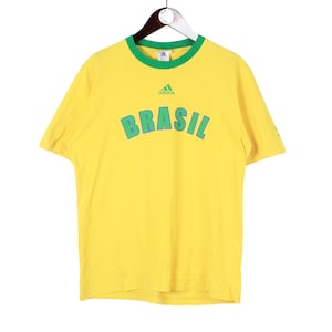 May include: Yellow t-shirt with green trim and the word "BRASIL" in green block letters. The Adidas logo is above the word. The shirt is hanging on a hanger.