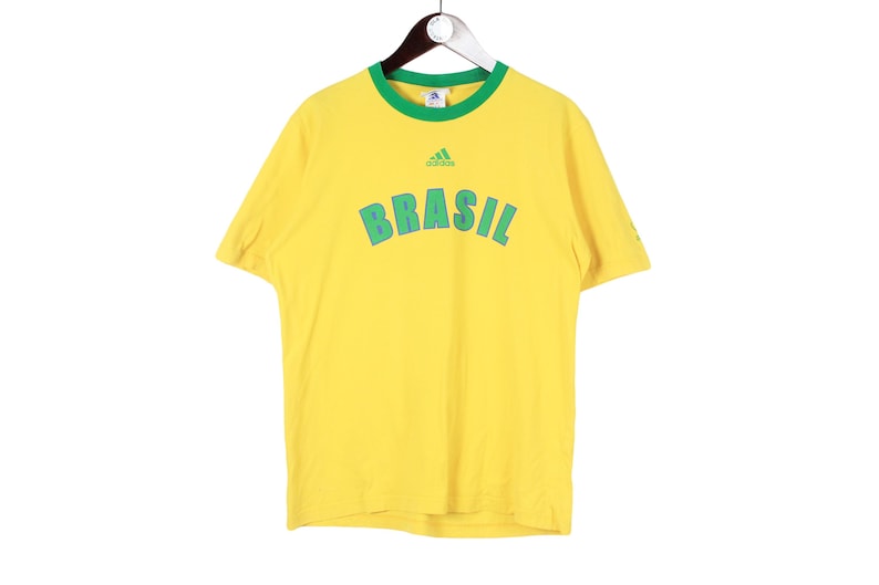 May include: Yellow t-shirt with green trim and the word "BRASIL" in green block letters. The Adidas logo is above the word. The shirt is hanging on a hanger.