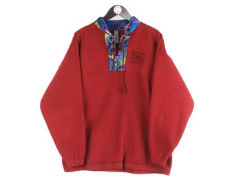 May include: A red fleece pullover with a patterned collar and a drawstring neckline. The text "BLUE ZONE OCEAN" is embroidered on the chest. The long-sleeved top is hanging on a black hanger.