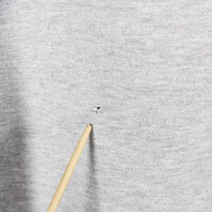 Puede incluir: Primer plano de una tela gris claro con un peque&ntilde;o agujero y un palillo de madera. La tela tiene un aspecto texturizado. El palillo est&aacute; inclinado hacia el agujero.