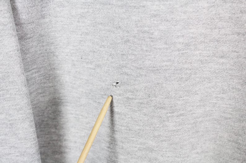 Puede incluir: Primer plano de una tela gris claro con un peque&ntilde;o agujero y un palillo de madera. La tela tiene un aspecto texturizado. El palillo est&aacute; inclinado hacia el agujero.