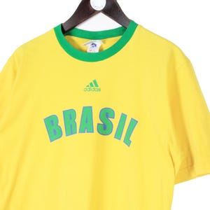 May include: Yellow t-shirt with green trim and the word "BRASIL" in large, blue letters. The Adidas logo is above the word "BRASIL". The shirt is hanging on a wooden hanger.