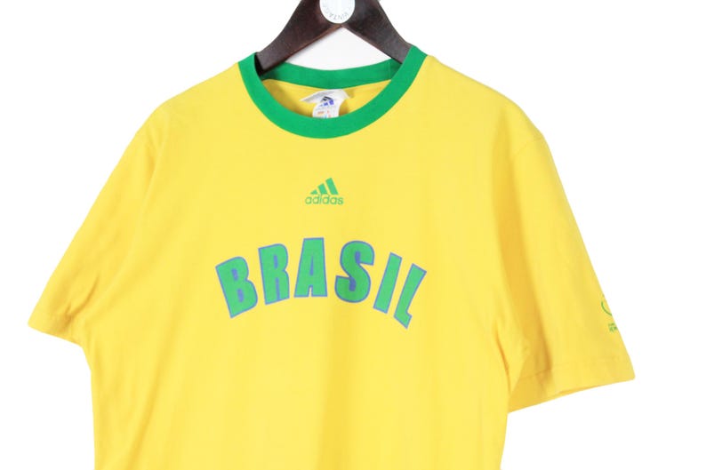 May include: Yellow t-shirt with green trim and the word "BRASIL" in large, blue letters. The Adidas logo is above the word "BRASIL". The shirt is hanging on a wooden hanger.