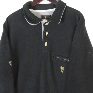 May include: Black fleece pullover with a collared neckline and three button closure. Features a small zippered pocket and a gold embroidered logo on the chest and sleeve. The collar is trimmed with white fabric.