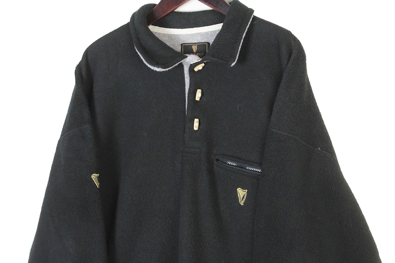 May include: Black fleece pullover with a collared neckline and three button closure. Features a small zippered pocket and a gold embroidered logo on the chest and sleeve. The collar is trimmed with white fabric.