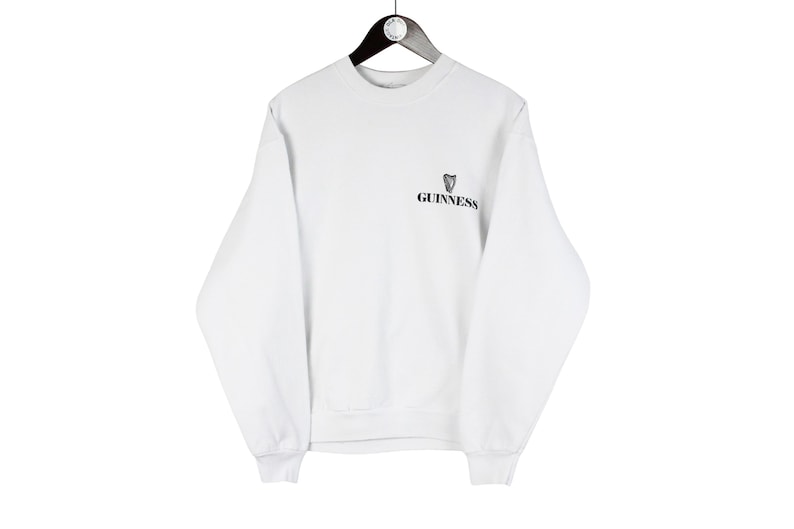 May include: White sweatshirt with the Guinness logo on the upper left chest. The long-sleeved crew neck shirt is hanging on a black hanger against a white background. The Guinness logo features a black harp and the Guinness wordmark.