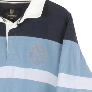 May include: A long-sleeved rugby shirt with a color-block design. The shirt features navy blue, light blue, and white horizontal stripes. A Guinness logo is embroidered on the chest. The collar is white.