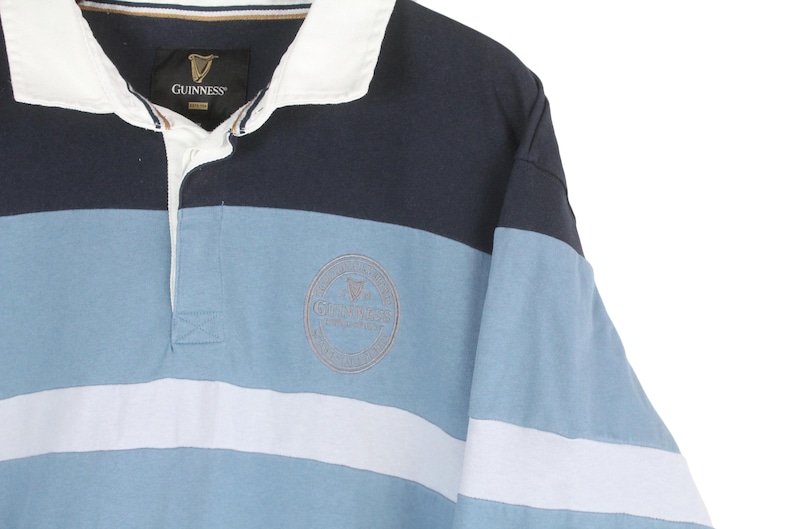 May include: A long-sleeved rugby shirt with a color-block design. The shirt features navy blue, light blue, and white horizontal stripes. A Guinness logo is embroidered on the chest. The collar is white.