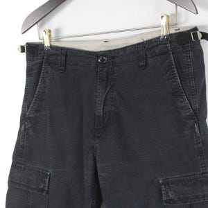 Puede incluir: Pantalones cortos cargo de color gris oscuro con m&uacute;ltiples bolsillos y cierre de bot&oacute;n. Los pantalones cortos est&aacute;n colgados de una percha de madera. La tela parece ser un material duradero y texturizado.