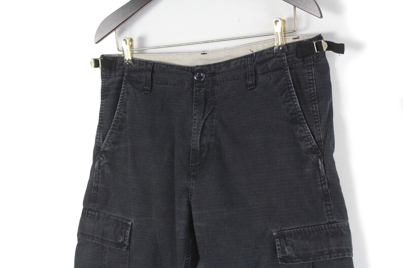 Puede incluir: Pantalones cortos cargo de color gris oscuro con m&uacute;ltiples bolsillos y cierre de bot&oacute;n. Los pantalones cortos est&aacute;n colgados de una percha de madera. La tela parece ser un material duradero y texturizado.