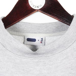 Puede incluir: Primer plano de una sudadera FILA gris con cuello redondo, colgada de una percha de madera. La etiqueta FILA es visible en el interior del cuello. La sudadera es de color gris claro.