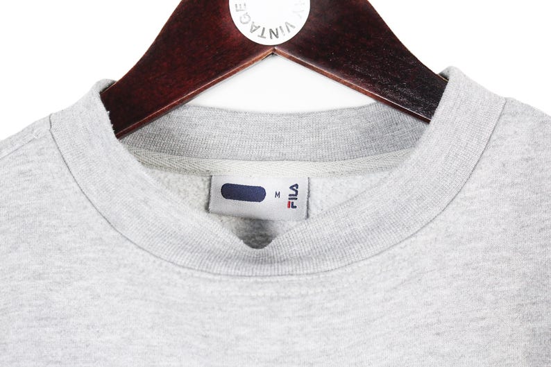 Puede incluir: Primer plano de una sudadera FILA gris con cuello redondo, colgada de una percha de madera. La etiqueta FILA es visible en el interior del cuello. La sudadera es de color gris claro.