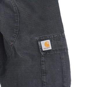 Puede incluir: Primer plano de un pantal&oacute;n cargo Carhartt gris oscuro. El pantal&oacute;n tiene una tela texturizada y un bolsillo rectangular en el lateral. Un parche con el logo de Carhartt con una 'C' naranja es visible en el bolsillo.