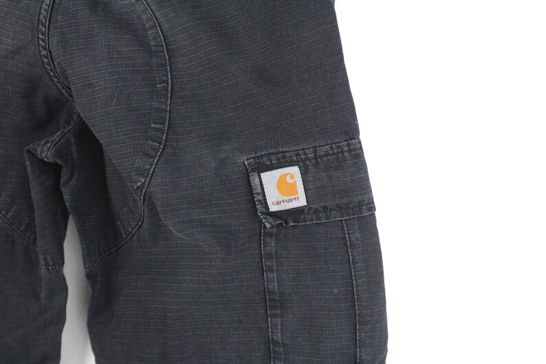 Puede incluir: Primer plano de un pantal&oacute;n cargo Carhartt gris oscuro. El pantal&oacute;n tiene una tela texturizada y un bolsillo rectangular en el lateral. Un parche con el logo de Carhartt con una 'C' naranja es visible en el bolsillo.