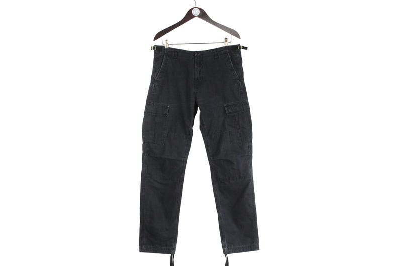 Puede incluir: Pantalones cargo negros colgados de una percha de madera. Los pantalones tienen m&uacute;ltiples bolsillos, cierre de bot&oacute;n y cremallera, y cordones en los tobillos. Los pantalones est&aacute;n hechos de una tela duradera.