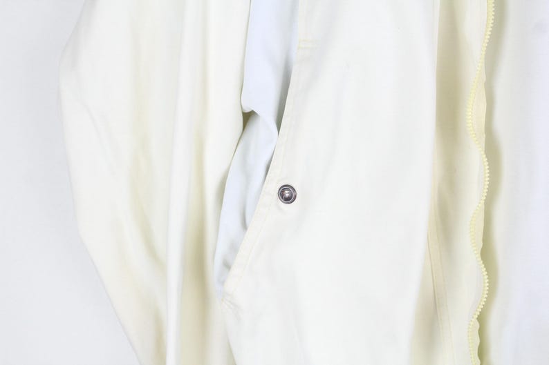 May include: A cream-colored jacket featuring a vertical zipper closure along the right side, a single visible metallic snap button on the front placket, and smooth fabric construction with clean, minimalist lines and a structured collar area.