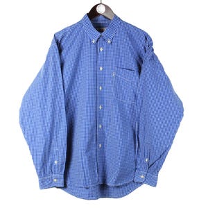 Può includere: Camicia a quadri blu e bianca con maniche lunghe, colletto button-down e taschino sul petto. La camicia è appesa a una gruccia di legno su sfondo bianco.