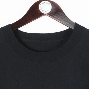 Op de afbeelding: Een zwart T-shirt met ronde hals hangt aan een houten kledinghanger. Het shirt is gemaakt van een effen zwart materiaal en heeft een klassiek, eenvoudig ontwerp. De hanger heeft een witte sticker met tekst.