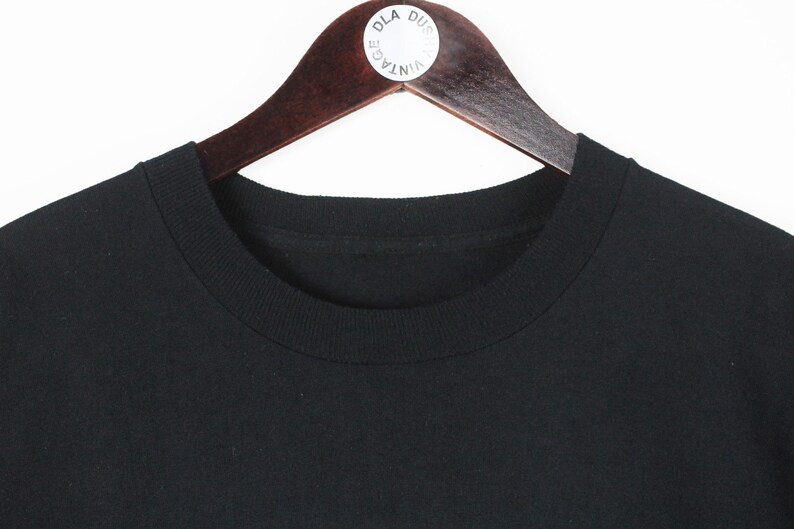 Op de afbeelding: Een zwart T-shirt met ronde hals hangt aan een houten kledinghanger. Het shirt is gemaakt van een effen zwart materiaal en heeft een klassiek, eenvoudig ontwerp. De hanger heeft een witte sticker met tekst.